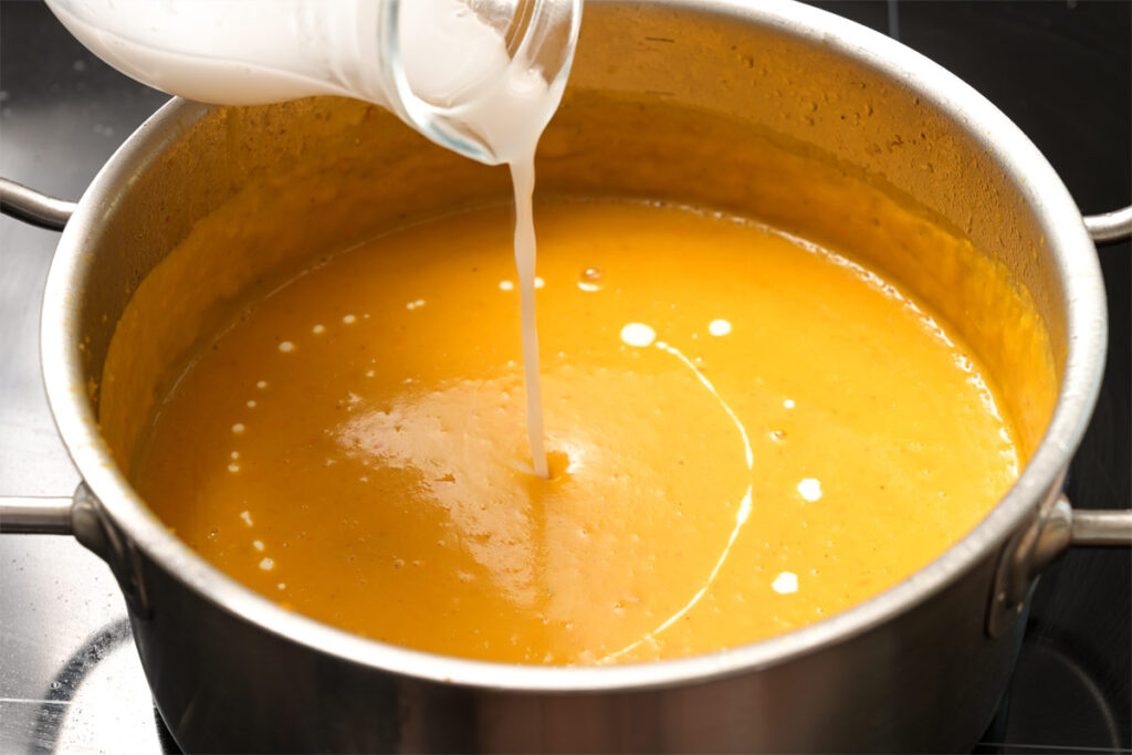 Frau gießt Kokosmilch in eine Gemüsecremesuppe im Stahltopf