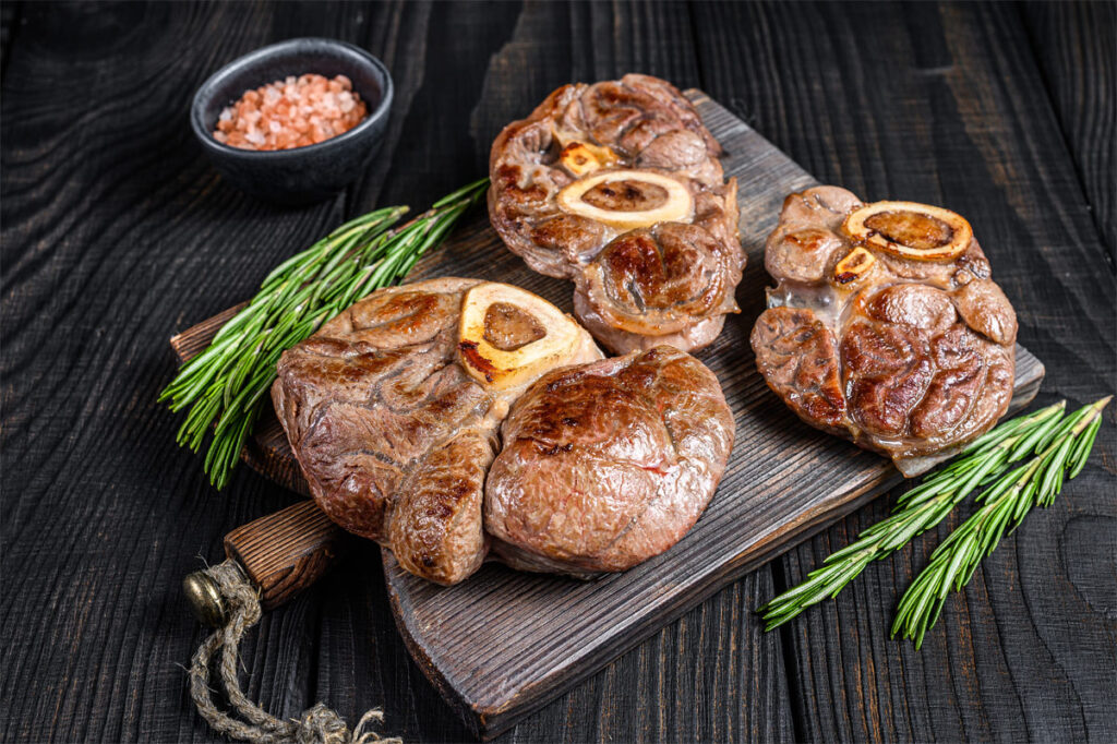 Geschmorte Kalbshaxe Ossobuco auf einem dunklen Holzbrett. Mit frischen Kräutern umlegt.