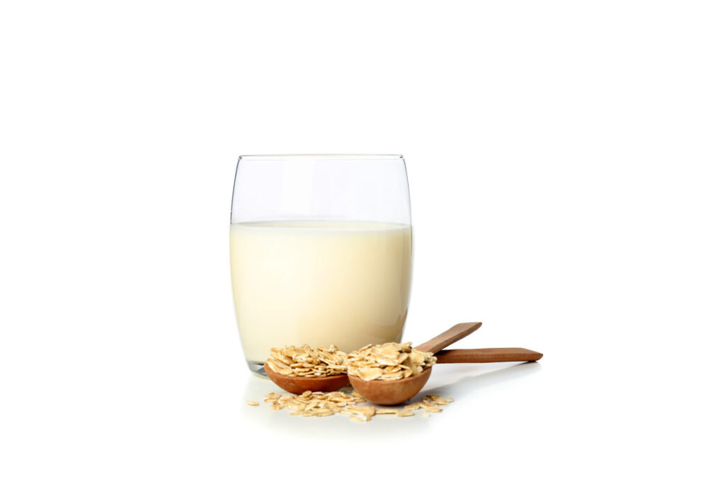 Ein Glas mit Hafermilch und zwei davorliegenden Holzlöffeln mit Haferflocken.